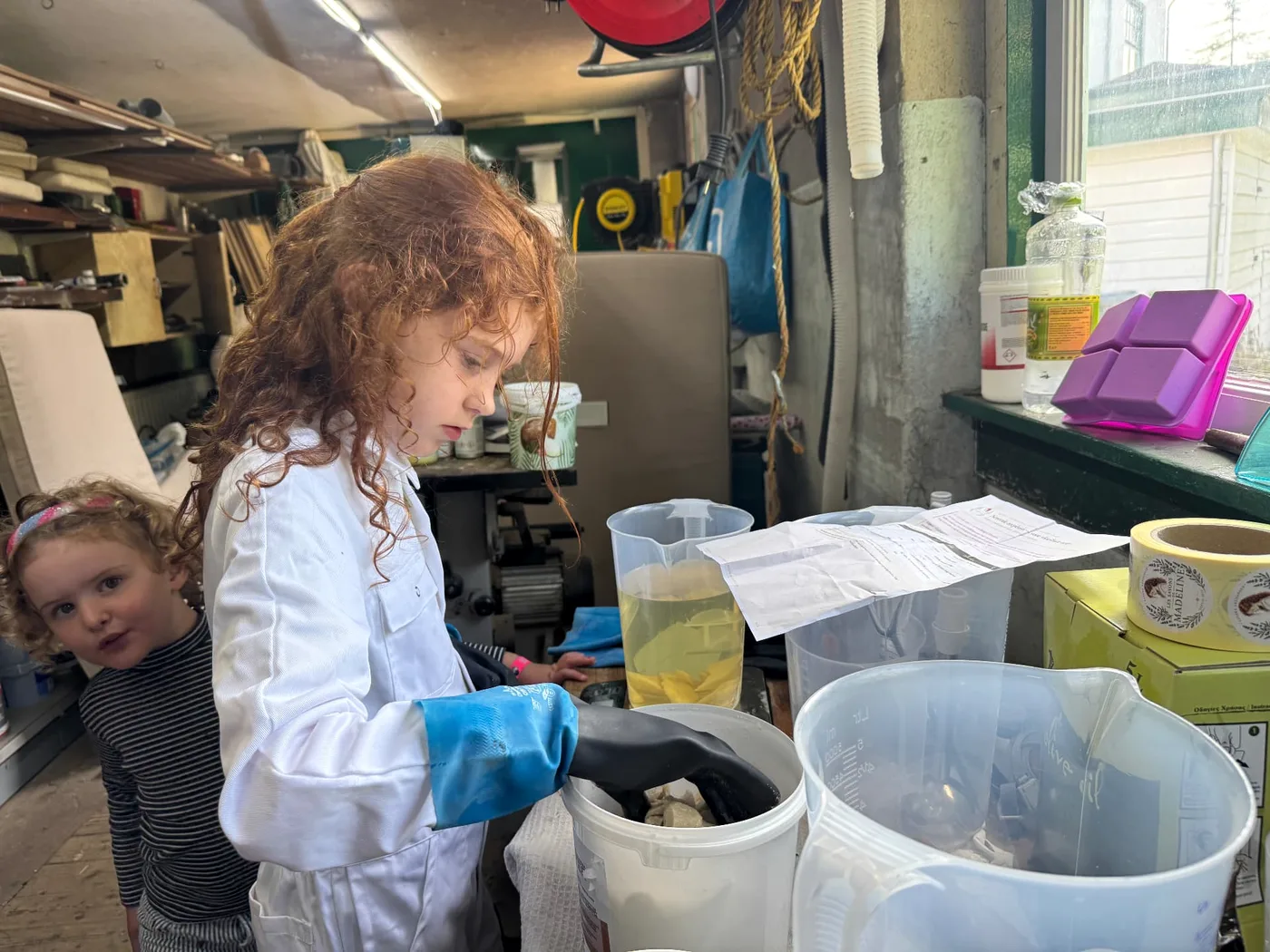 Madeline in werkjas en handschoenen bij de maatbekers met olijfolie in het atelier